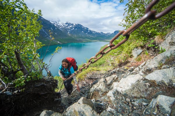 En turgåer med sekk holder seg fast i en sikkerhetskjete på en bratt, steinete fjellsti, med en vakker turkis innsjø og snøkledde fjell i bakgrunnen.