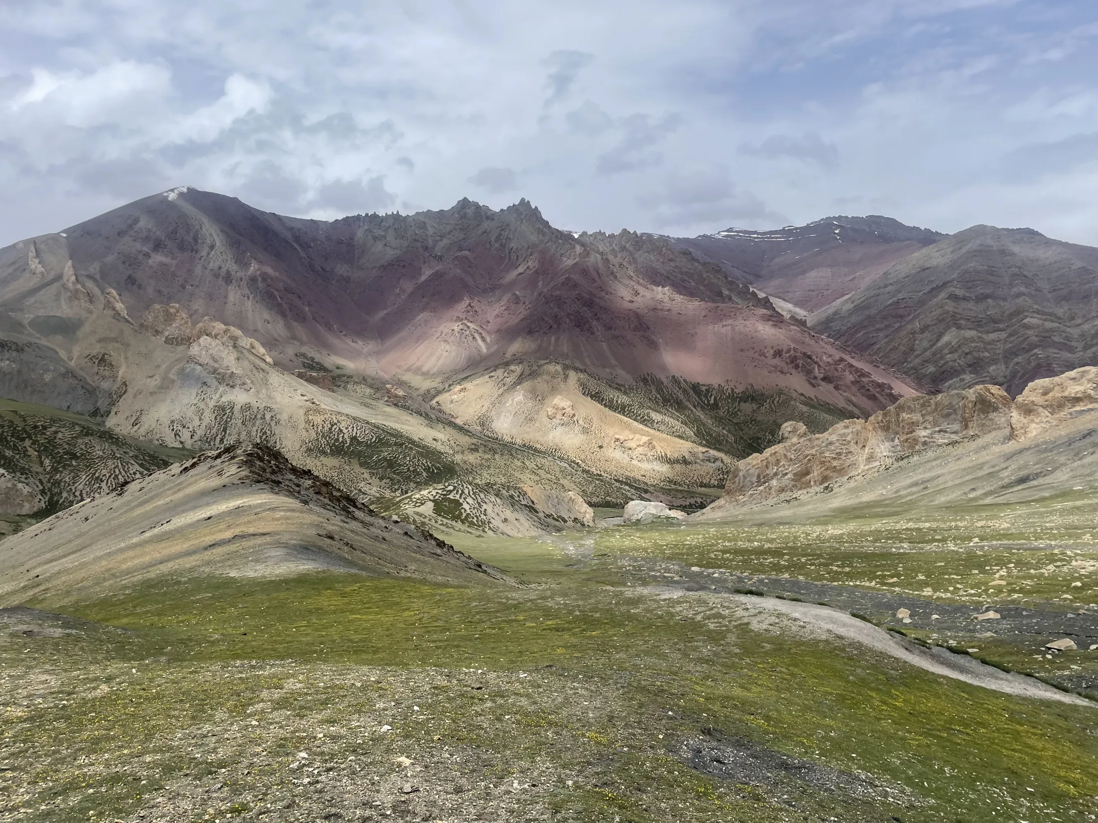 Et vidstrakte fjellandskap viser grønne, bølgende skråninger i forgrunnen som fører mot en dyp dal og dramatiske, flerfargede fjelltopper i rød-lilla og beige toner under en skyet himmel.