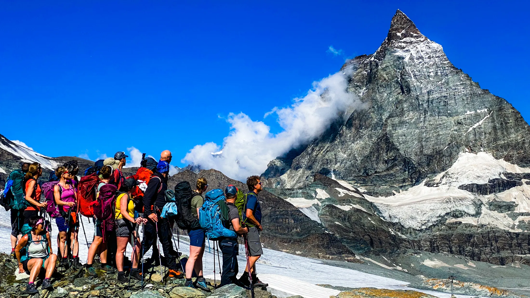 En gruppe turgåere med ryggsekker står på et steinete utsiktspunkt og ser opp på den ruvende, snødekte toppen av Matterhorn mot en klar, blå himmel.