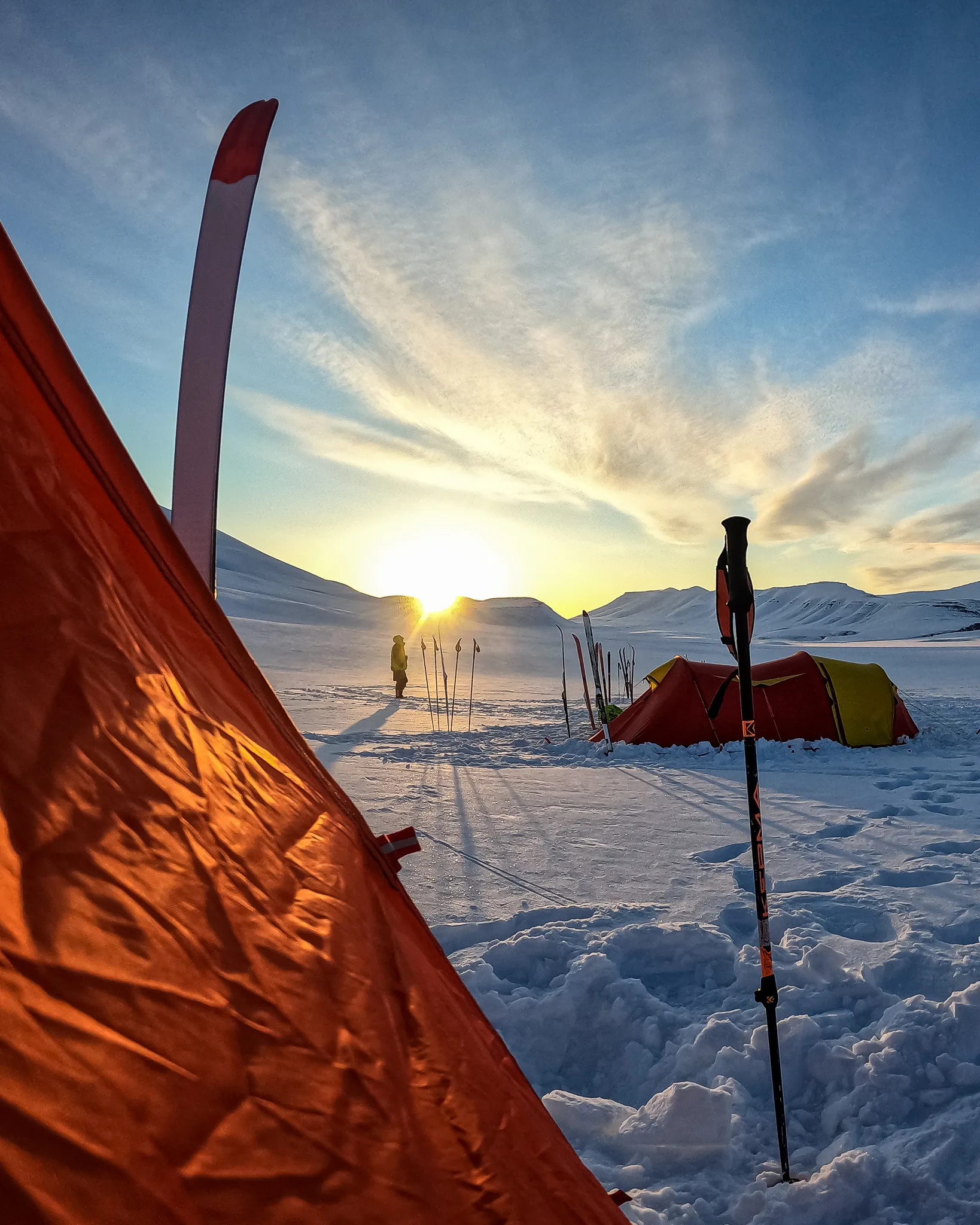 En arktisk teltleir med oransje telt satt opp på snødekt mark mens en enslig person står og ser solen gå lavt ned mellom fjerne fjell.