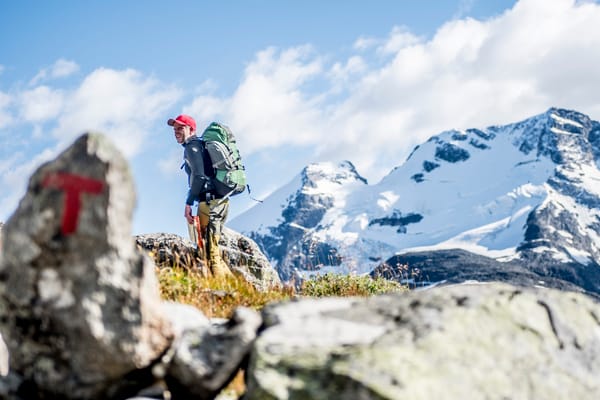En turgåer med stor sekk på en fjellsti ved en stein merket med rød T, med snødekte topper i bakgrunnen.