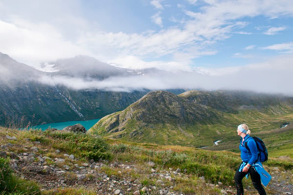 En turgåer med blå jakke og sekk går ned en gresskledd fjellsti mot et turkis vann under en skyet himmel.