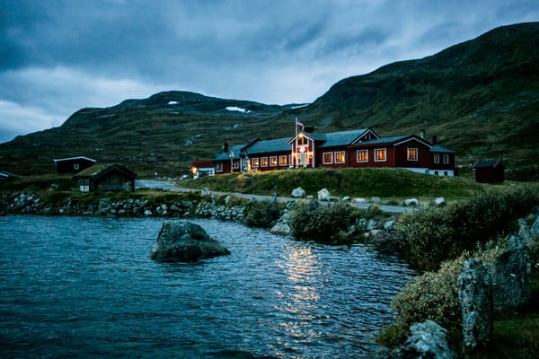 En rød fjellstue ved et mørkt vann under en blå kveldshimmel.