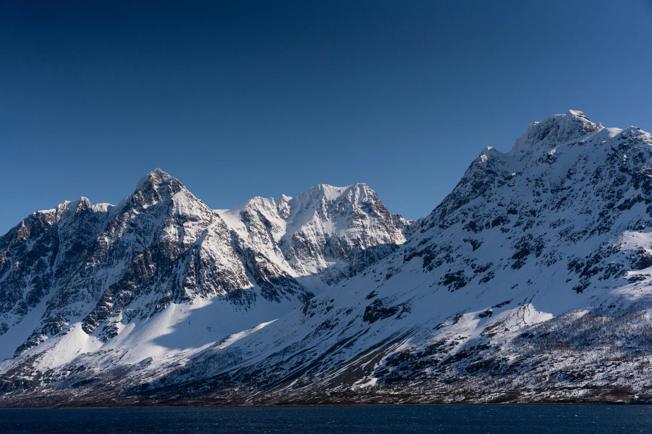 Snødekte fjell under en blå himmel ved sjøen.