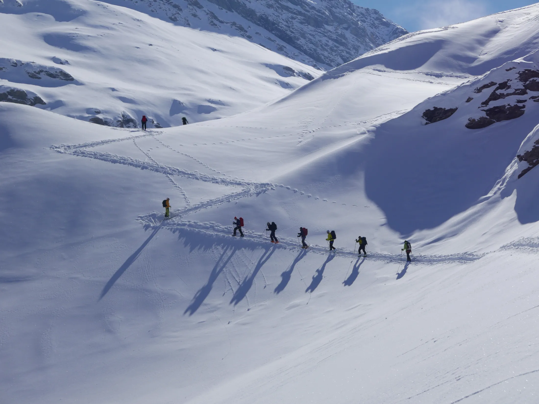 En rekke toppturvandrere går oppover en snødekt fjellside og kaster lange, dramatiske skygger over den urørte snøen mens de følger et sikksakk-spor.