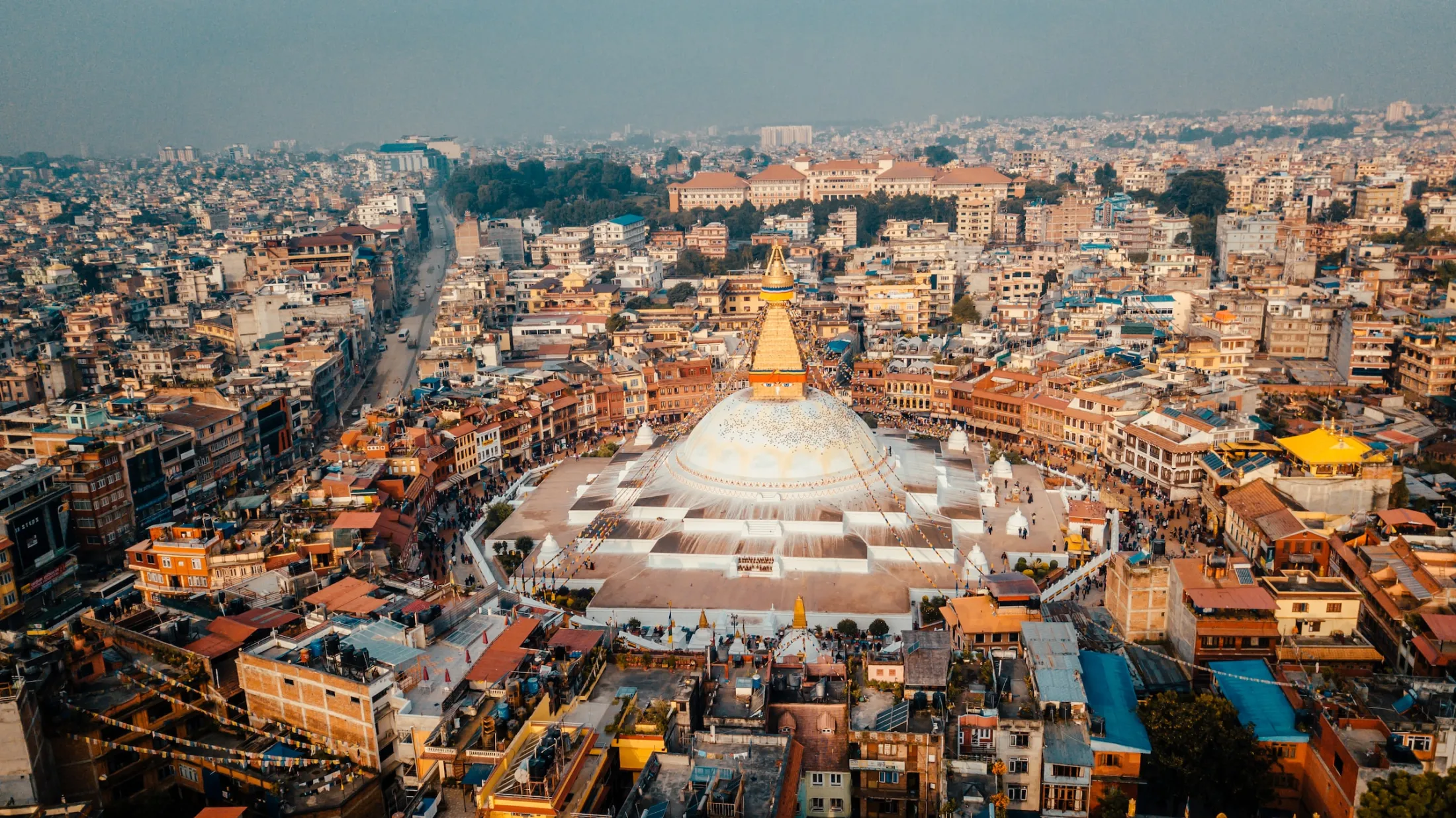 Et høytvinklet flyfoto viser den massive hvite domen og det gylne spiret til Boudhanath-stupaen sentrert på en sirkulær plass, helt omkranset av Katmandus tette bylandskap under en disig himmel.