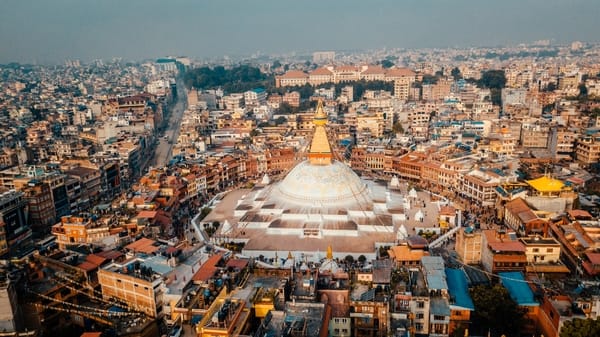 Et høytvinklet flyfoto viser den massive hvite domen og det gylne spiret til Boudhanath-stupaen sentrert på en sirkulær plass, helt omkranset av Katmandus tette bylandskap under en disig himmel.