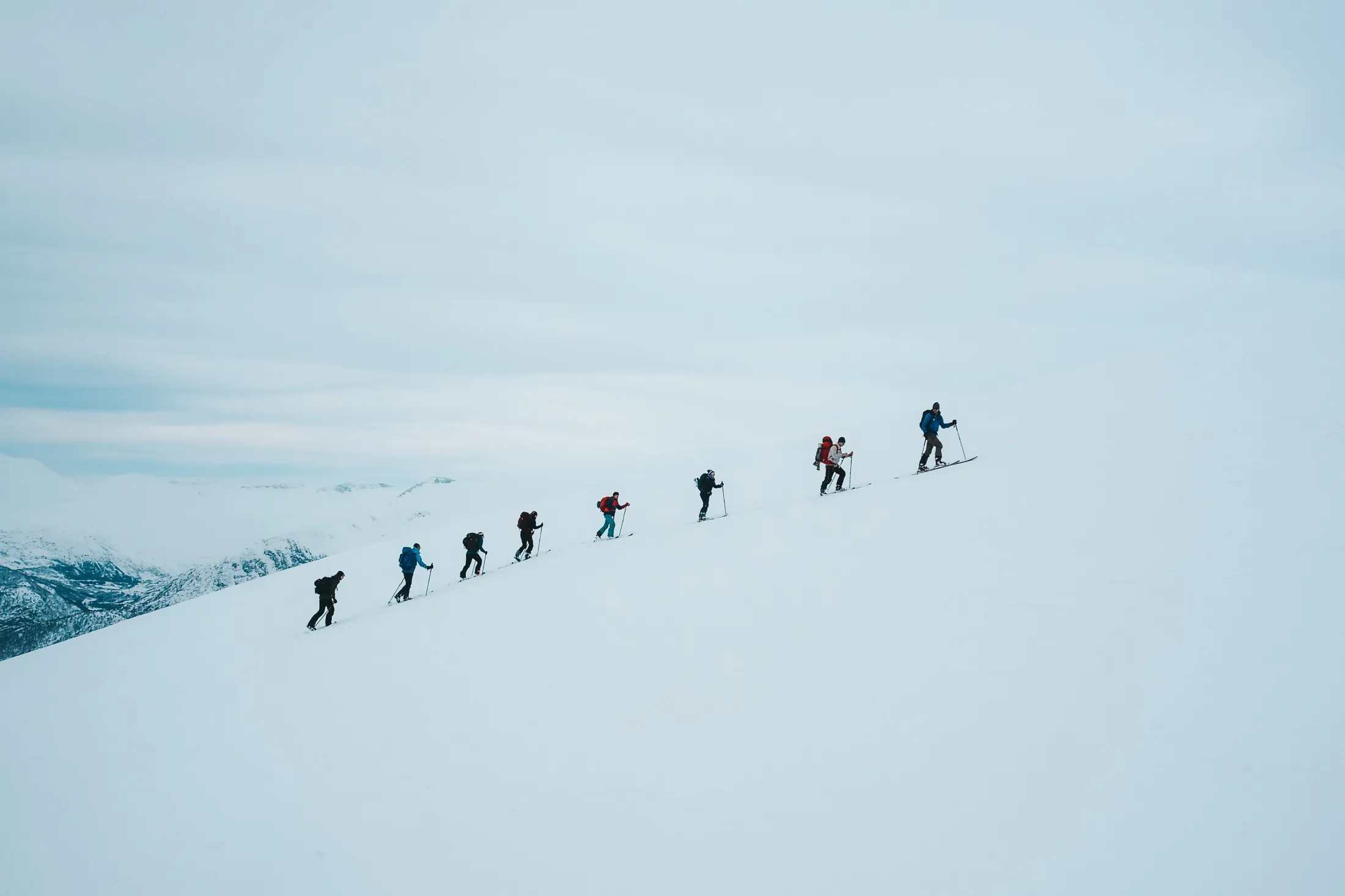 En gruppe skiløpere går på rekke oppover en bratt, snødekket fjellside under en overskyet himmel.