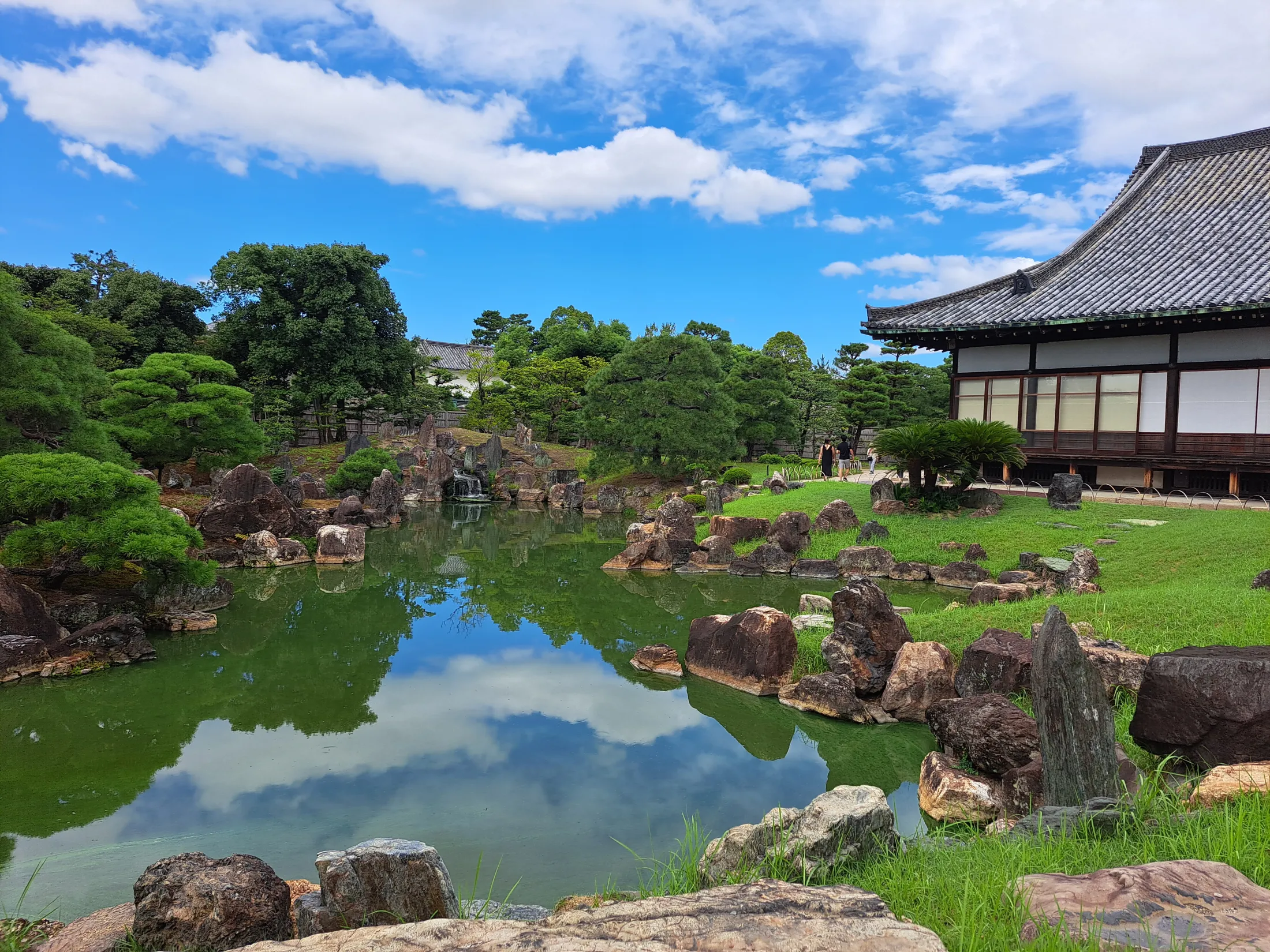 En fredelig tradisjonell japansk hage har en rolig dam som speiler en blå, skyet himmel, omkranset av nøyaktig plasserte steiner, frodige grønne trær og en historisk trebygning.
