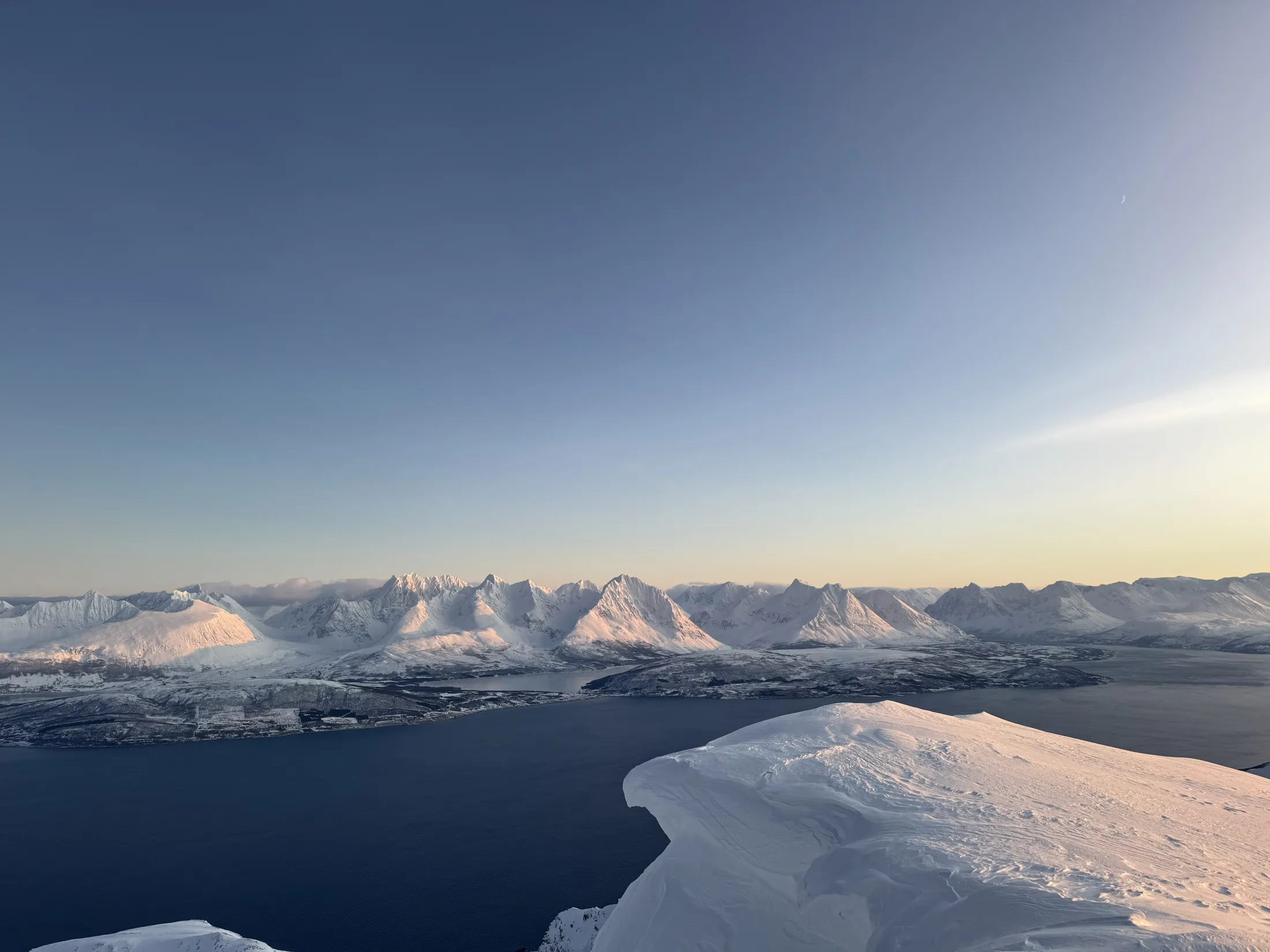 Flyfoto av en snødekket fjellrygg under en klar blå himmel, med utsikt over en mørkeblå fjord og en kystbygd nedenfor.