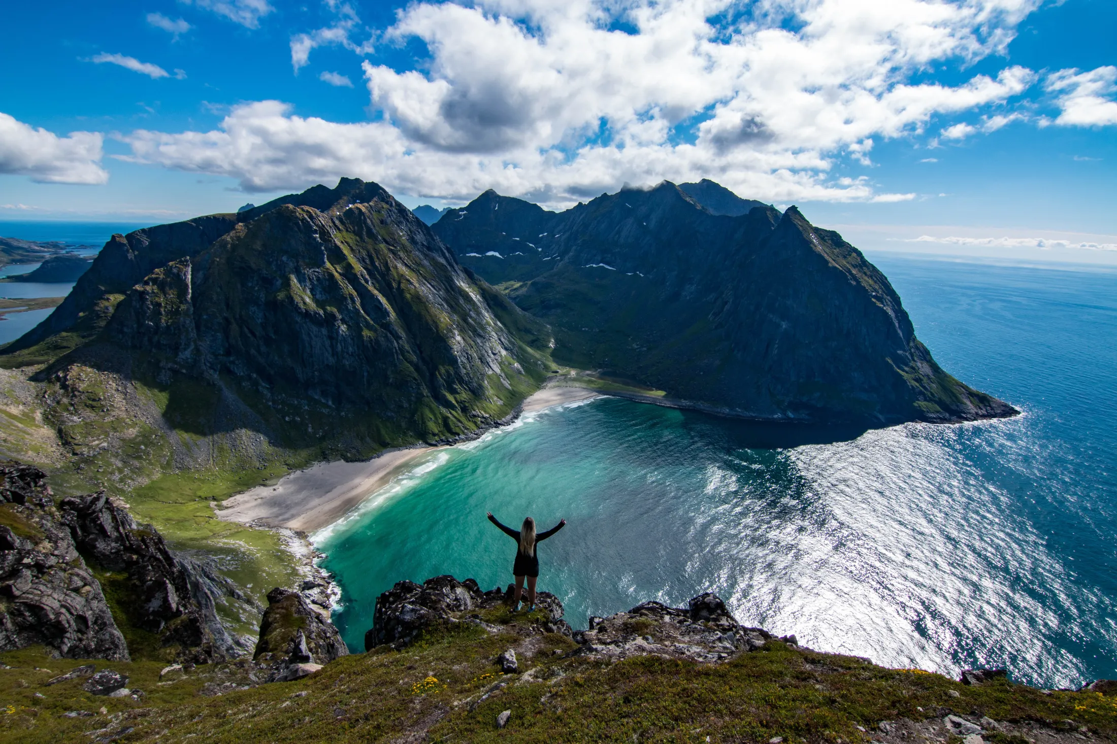 En turgåer med armene hevet i seier står på en fjelltopp med utsikt over en strand og havet nedenfor.