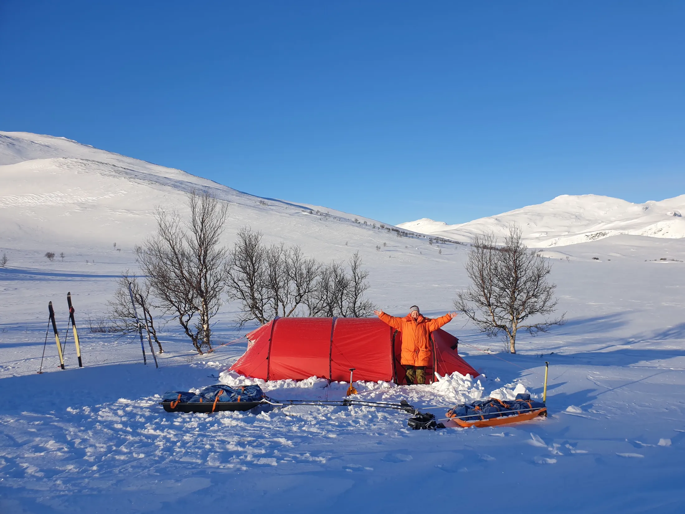 En person i oransje jakke står ved et rødt telt i et snødekt fjelllandskap under en blå himmel.