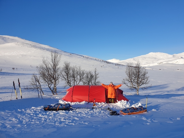 En person står foran et rødt telt i et snødekt fjelllandskap under en klar blå himmel.