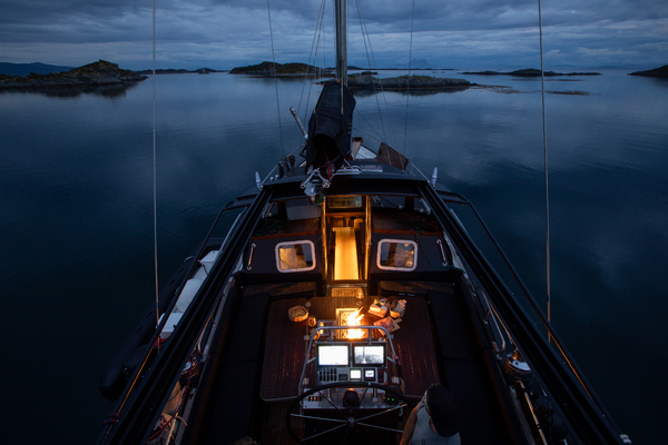 En seilbåt navigerer rolig, mørkt vann i skumringen, med øyer i bakgrunnen.