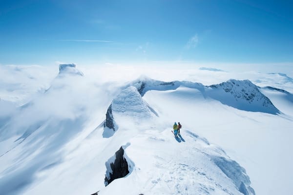 Luftfoto av to skiløpere på en snødekket fjellrygg under en klar blå himmel.