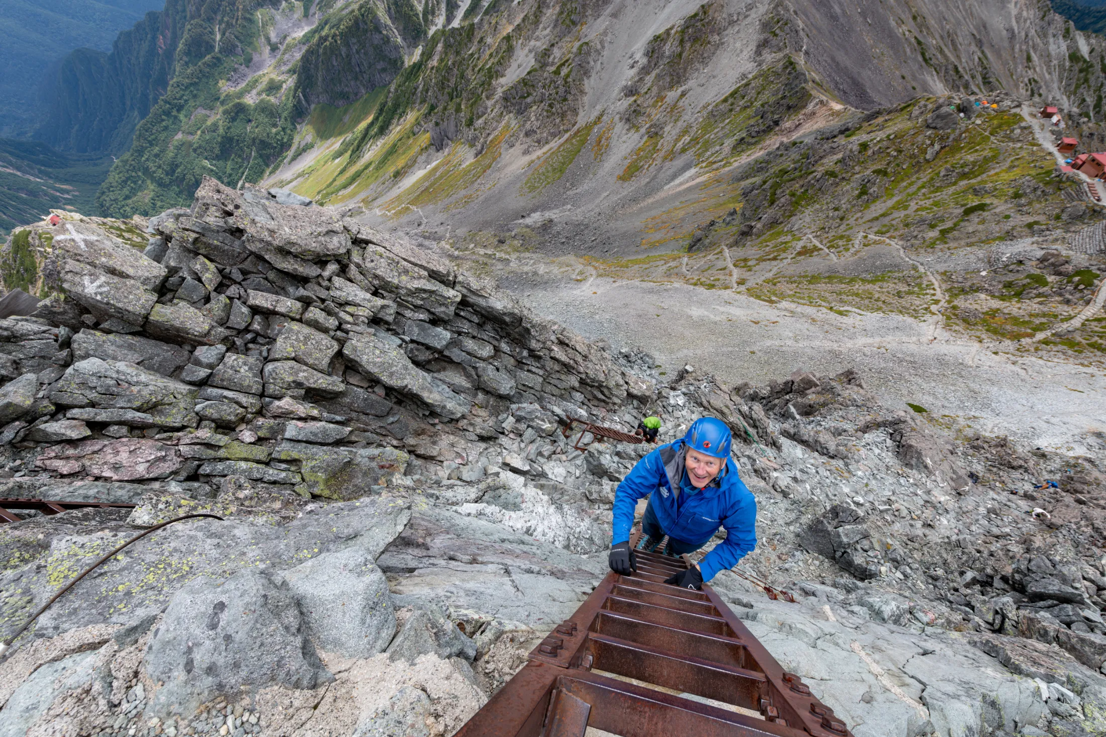Sett ovenfra ned en bratt, steinete fjellside, klatrer en smilende person i blå jakke og blå hjelm opp en lang metallstige som er boltet fast til klippen.