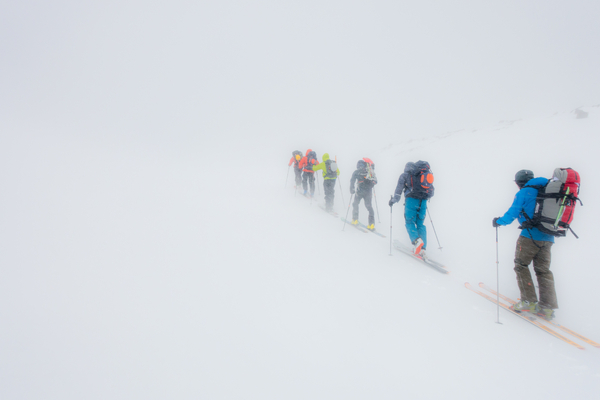 En gruppe skiløpere med sekk går opp et snødekt fjell i tett tåke.