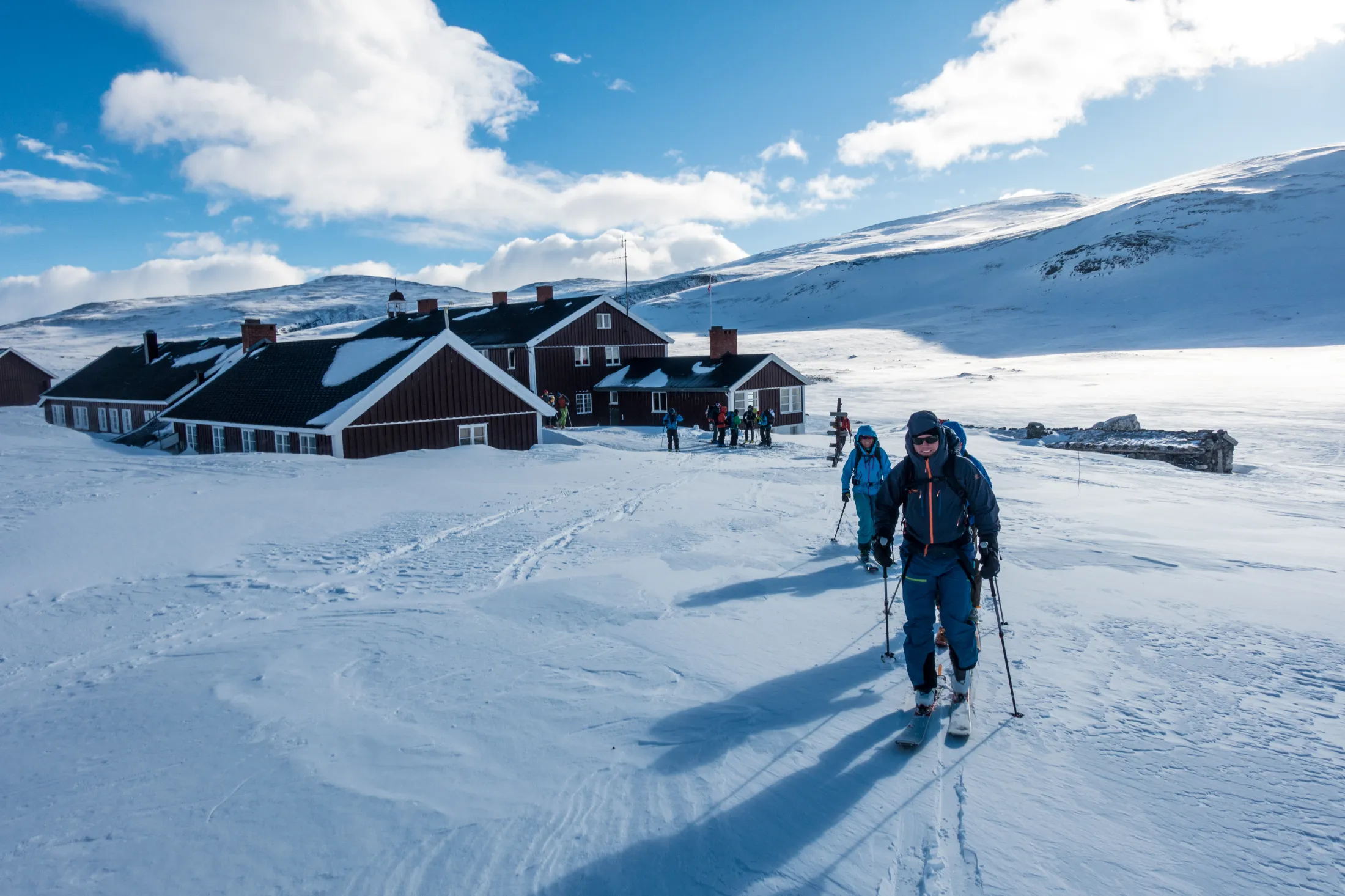 Personer som går på ski mot en rød tømmerhytte i et snølandskap under blå himmel.