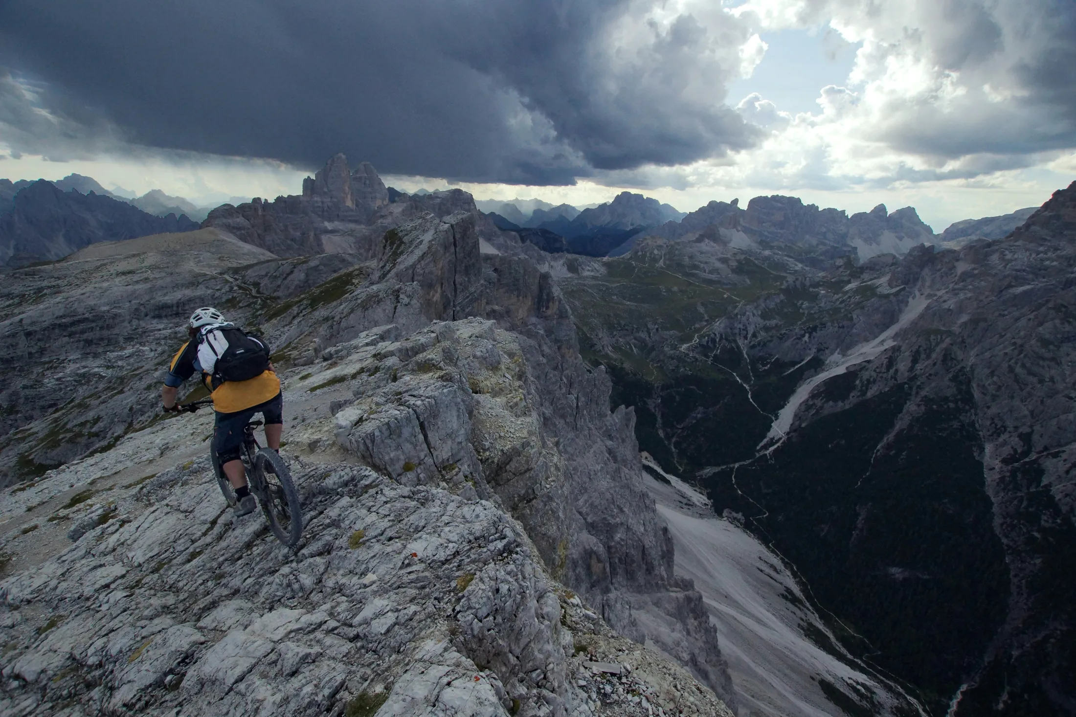 En terrengsyklist som sykler langs en smal, steinete fjellrygg under en dramatisk, overskyet himmel i Alpene.