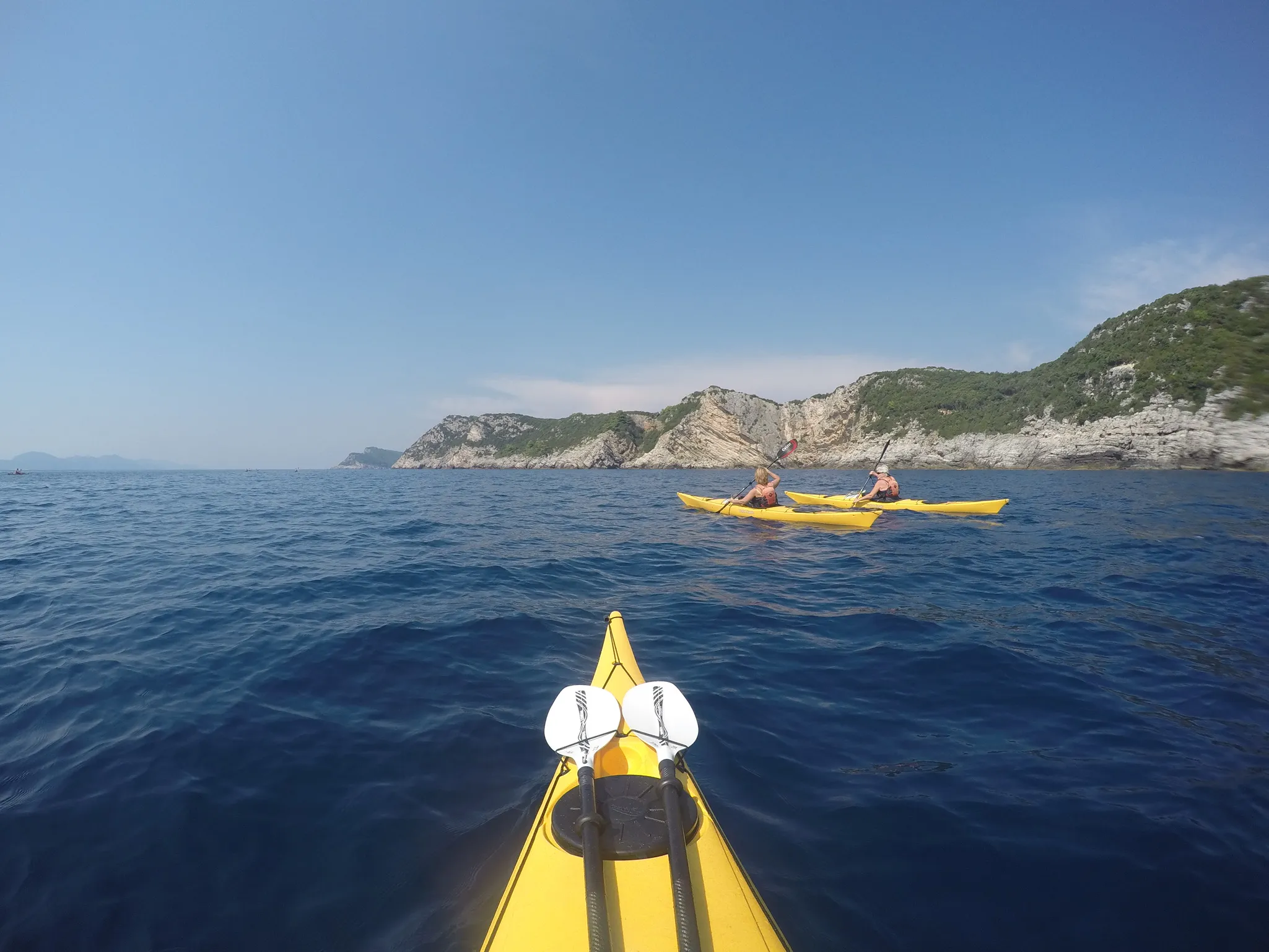 En padlers perspektiv fra kajakken på havet, med utsikt mot en lang kystlinje preget av klipper og frodig vegetasjon.