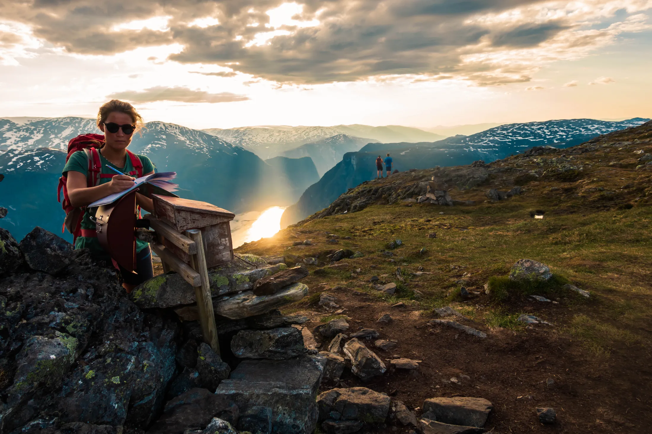 Dame på en fjelltopp med solnedgang i bakgrunn.