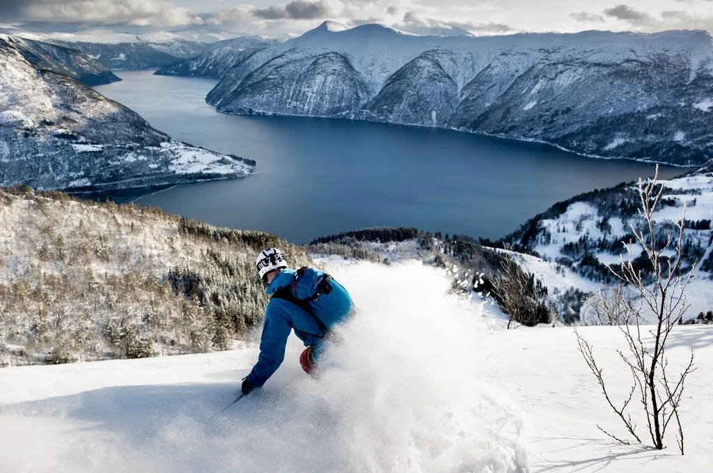 En skiløper i blå jakke svinger i løssnøen ned en fjellside med en dyp, blå fjord og snødekte fjell i bakgrunnen.
