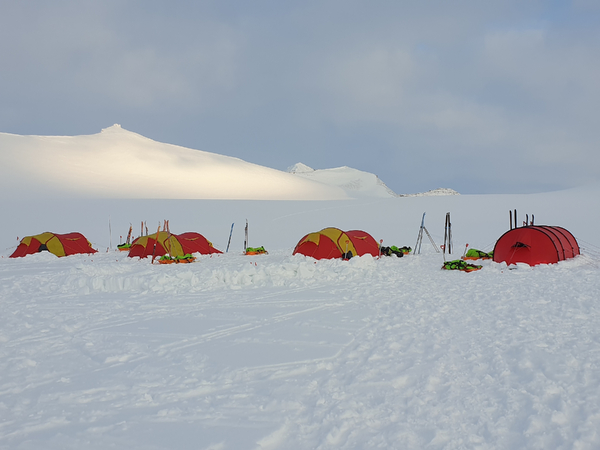 Flere røde og gule telt satt opp i et snølandskap med fjell i bakgrunnen under en overskyet himmel.