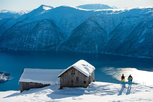 To skiløpere står ved siden av to snødekte trehytter og ser utover en dyp blå fjord omkranset av massive, snødekte fjell.
