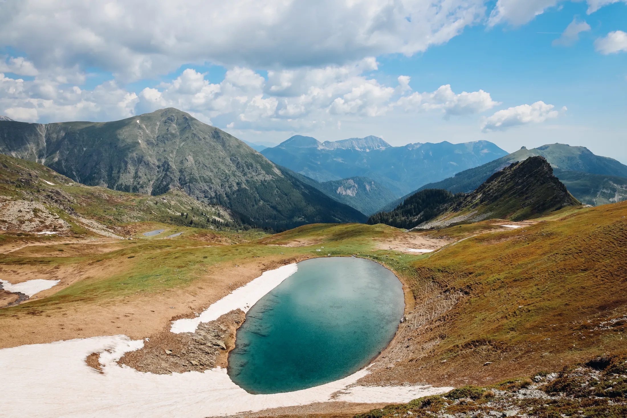 Et flybilde av et turkisfarget alpint vann omgitt av fjell og snøflekker under en delvis skyet himmel.