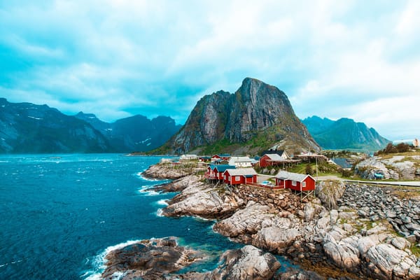 Røde rorbuer på en steinete kyst i Lofoten, med asurblått vann og dramatiske fjell under en lys himmel.