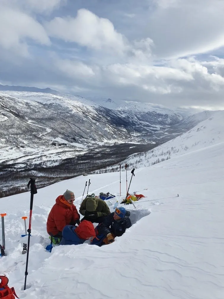 En gruppe mennesker som graver og arbeider i en snøprofil på en snødekt fjellside med utsikt over en dal i bakgrunnen.