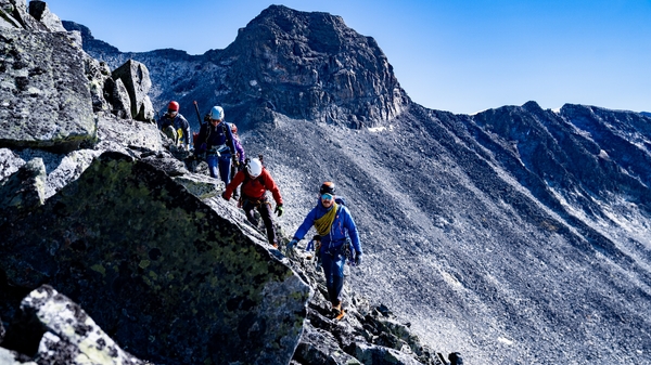 Fjellvandrere som klatrer opp en bratt og steinete fjellrygg under en klar blå himmel.