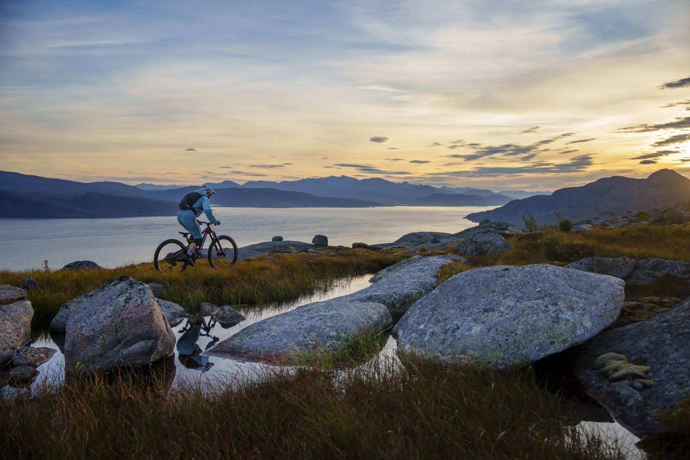 En terrengsyklist på sti med utsikt over ved sjøen og fjell i solnedgang.