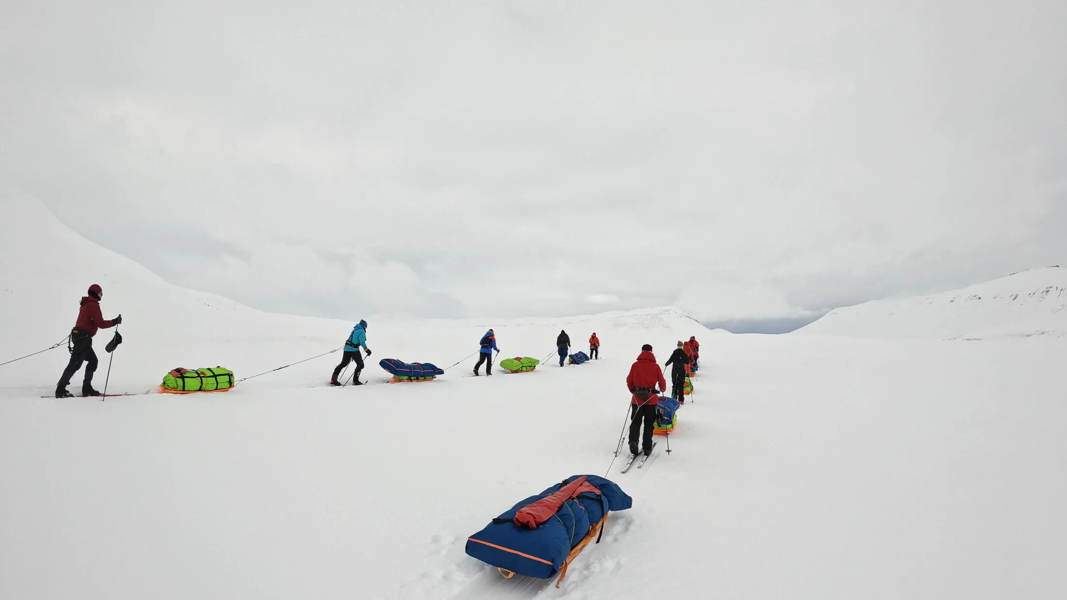 En gruppe ekspedisjonsskiløpere trekker fargerike pulker over en hvit, snødekt dal under en overskyet grå himmel.