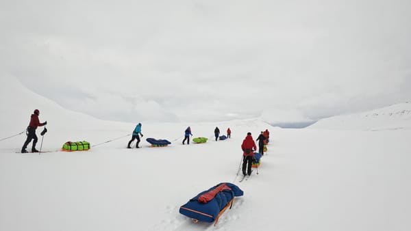 En gruppe ekspedisjonsskiløpere trekker fargerike pulker over en hvit, snødekt dal under en overskyet grå himmel.