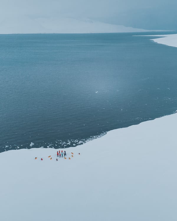 Et luftfoto av en liten ekspedisjonsgruppe som står i en sirkel med pulkene sine på kanten av en snødekt strand ved det kalde, åpne havet.