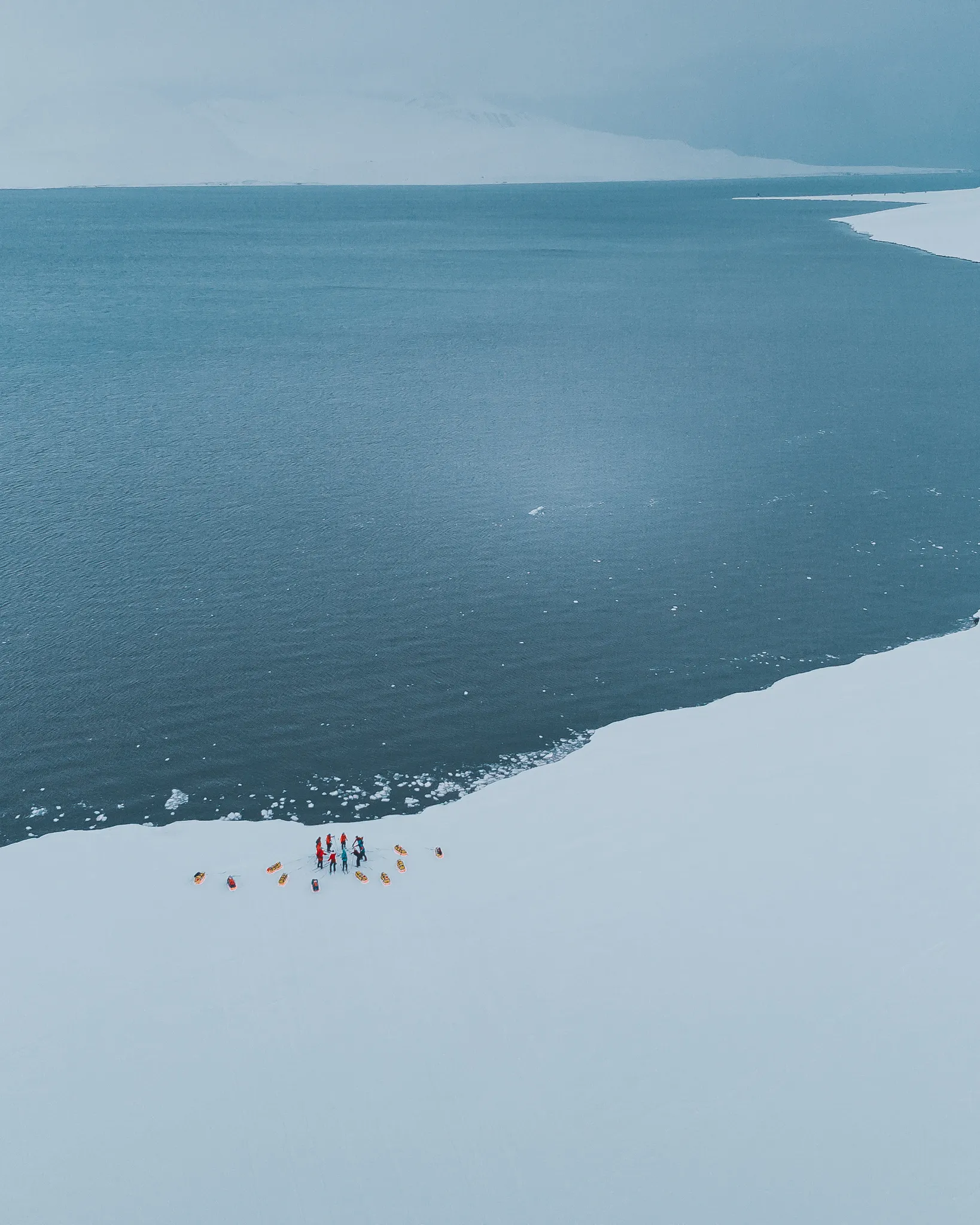 Et luftfoto av en liten ekspedisjonsgruppe som står i en sirkel med pulkene sine på kanten av en snødekt strand ved det kalde, åpne havet.