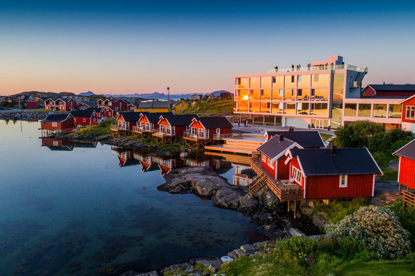Kystlandskap med tradisjonelle røde rorbuer og en moderne bygning langs strandkanten i solnedgang.