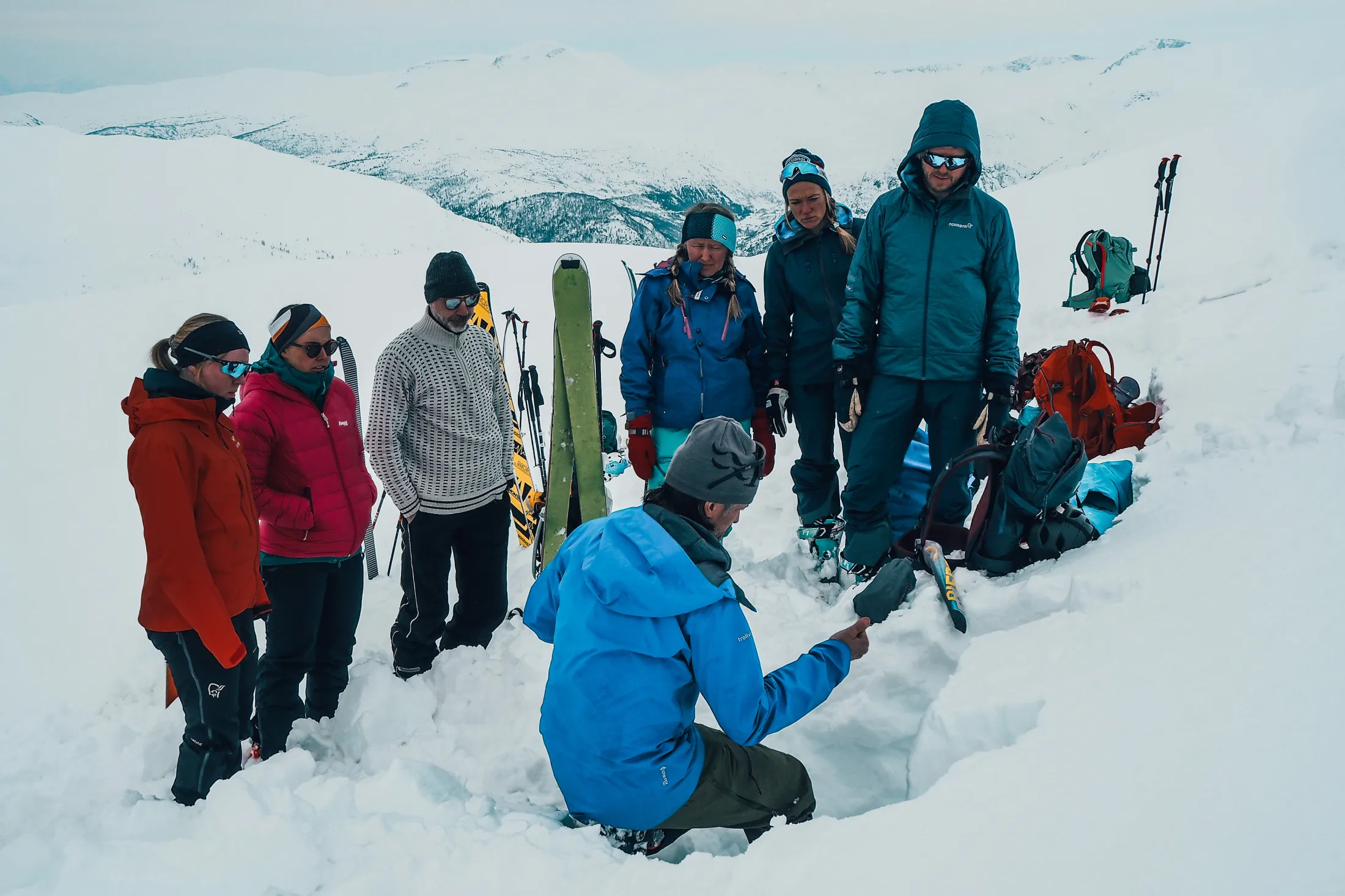 En guide i blå jakke demonstrerer snøforhold i en snøprofil for en gruppe skiturister i et snødekt fjellandskap.