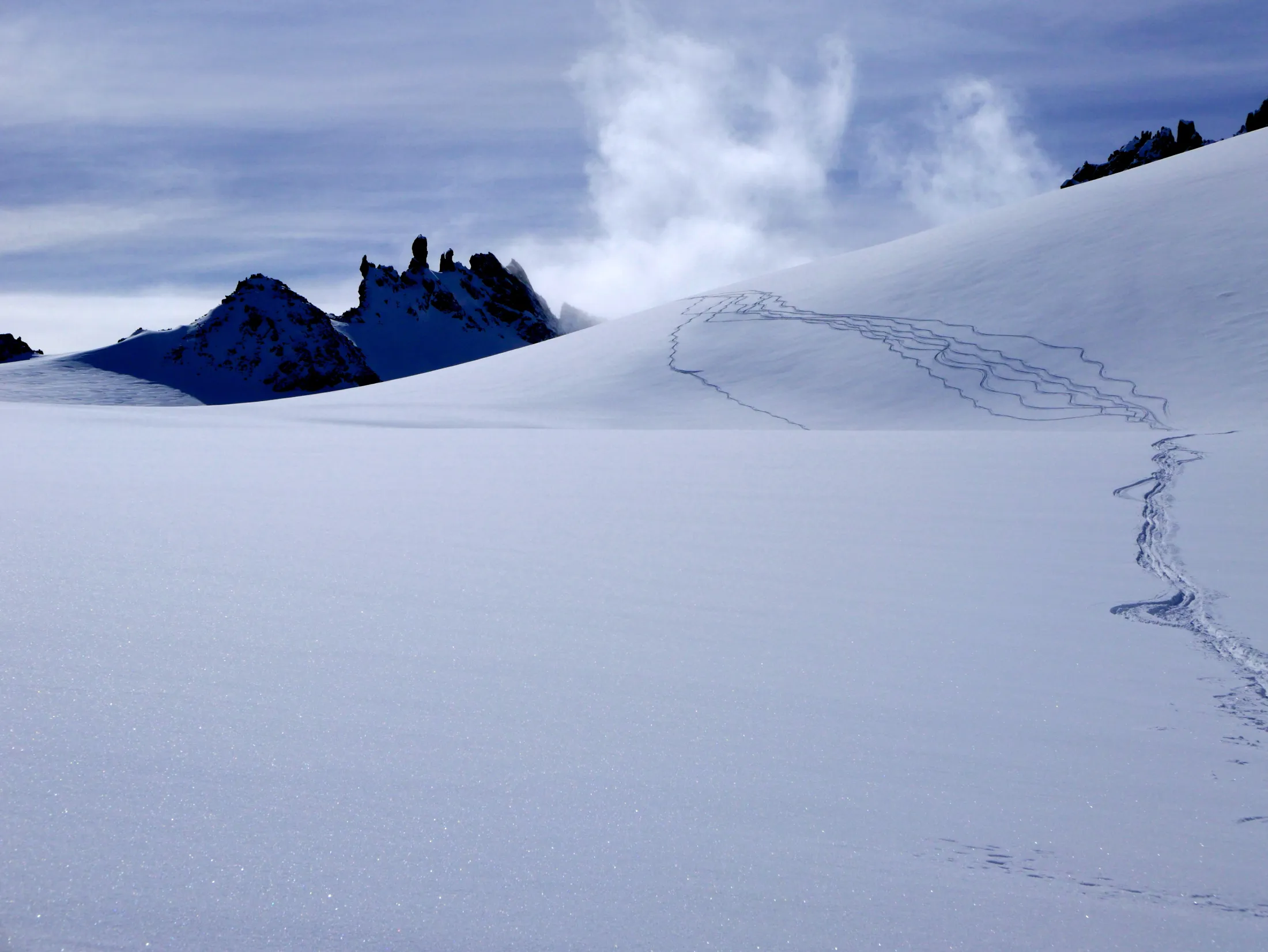 Et svingete skispor bukter seg over et enormt, urørt snøfelt som fører mot robuste fjelltopper delvis skjult av drivende skyer.