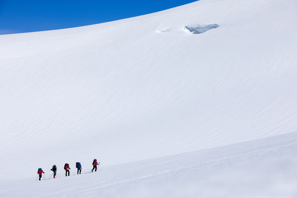 Fem personer går opp en stor, snødekt fjellskråning under en blå himmel.