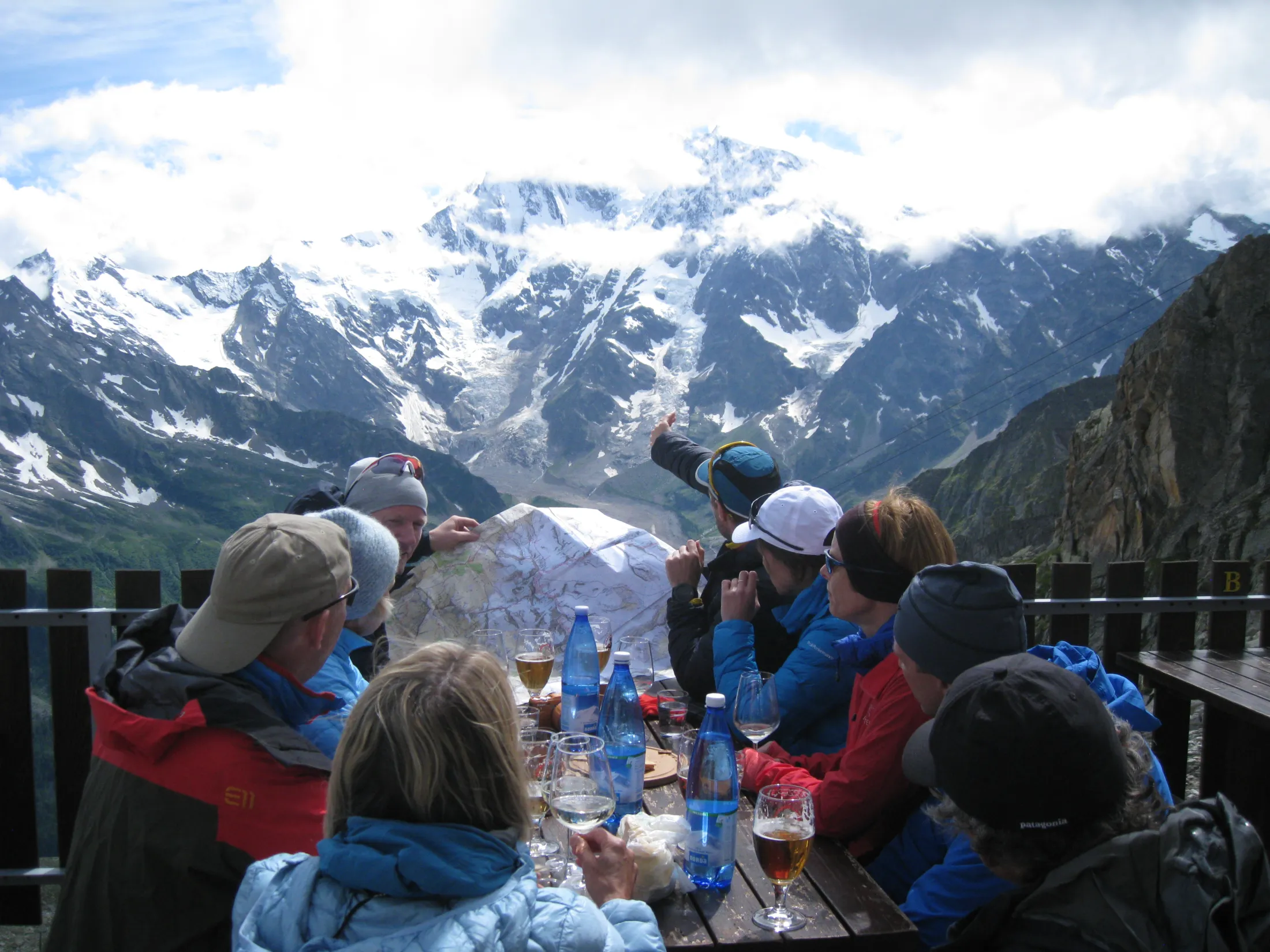 En gruppe turgåere samles rundt et utendørs trebord med drinker og et kart, og ser mot en massiv, snødekt fjellvegg delvis dekket av skyer.