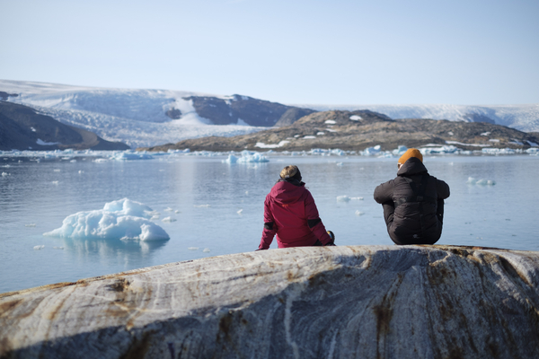 To personer sitter på et svaberg og skuer ut over en blå fjord fylt med lyseblå isfjell, med en stor isbre og snødekte fjell i bakgrunnen.