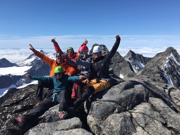 En gruppe fjellklatrere feirer på en fjelltopp med utstrakte armer mot en blå himmel.