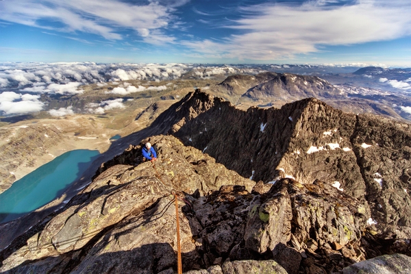 Utsikt ovenfra en fjelltopp, med utsikt over kuperte fjell og et turkis vann.