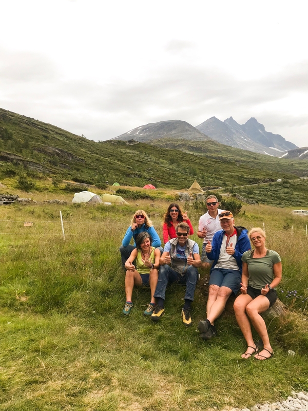 En gruppe på sju personer som slapper av i et gresskledd fjelllandskap med telt i bakgrunnen.