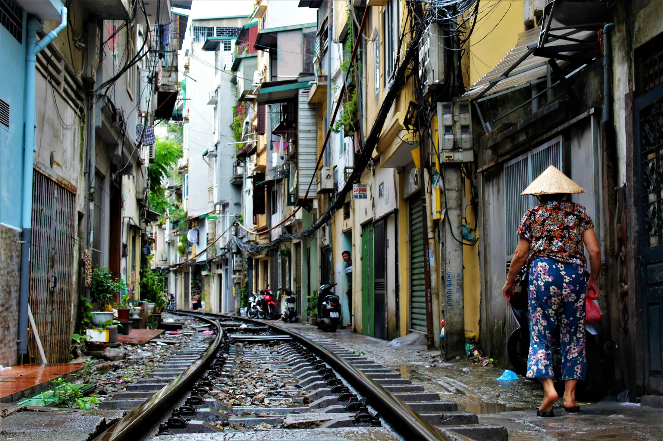 Ei kvinne med kjegleformet hatt går langs ei smal jernbanelinje flankert av tett i tett med bygninger i Hanoi.