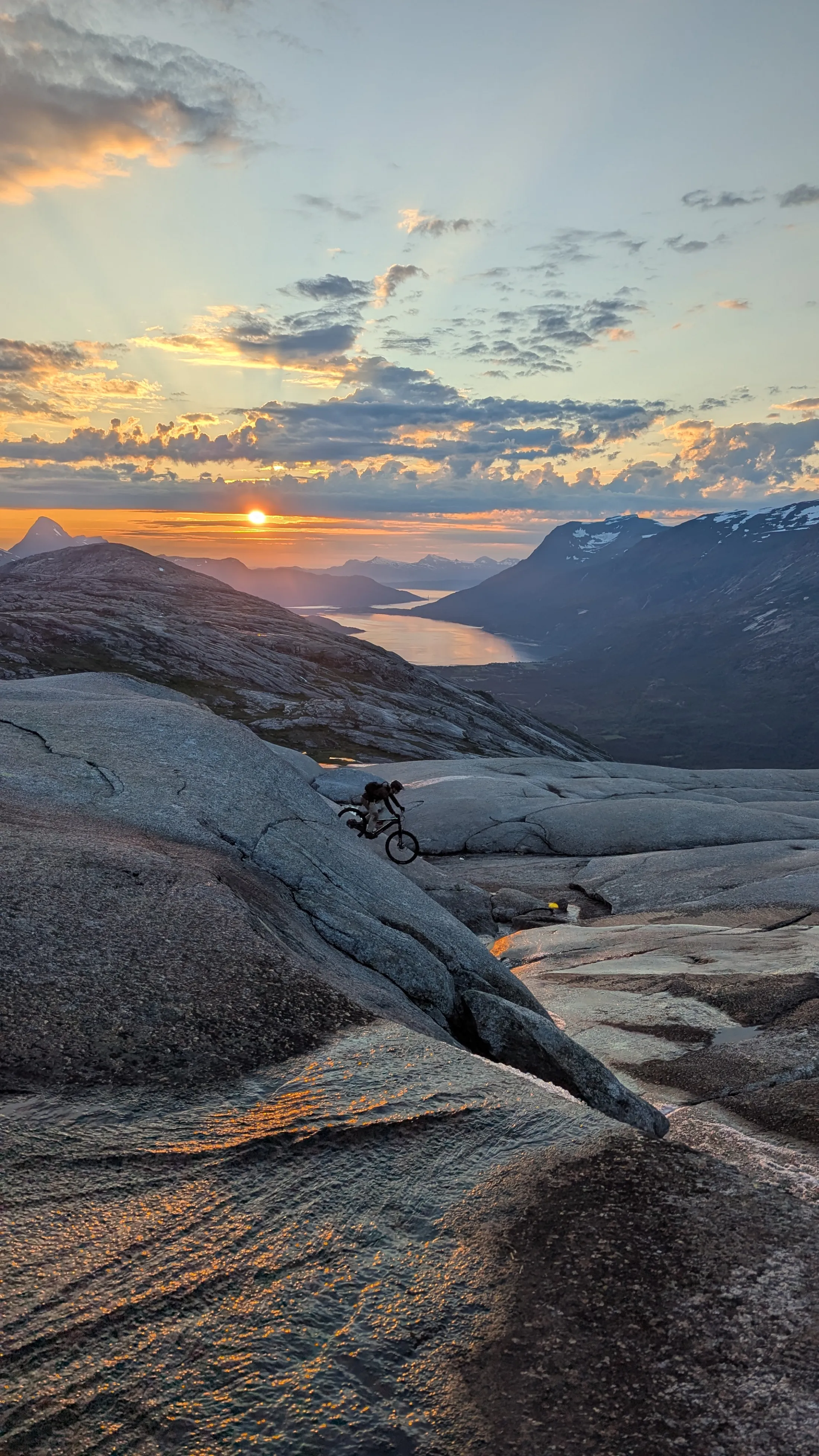 En terrengsyklist på glatt svaberg i silhuett mot en dramatisk solnedgang med fjerne fjell og en innsjø under.