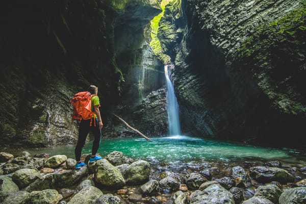 En turgåer med en knalloransje ryggsekk står på mosegrodde steiner og ser på en skjult foss som renner ned i et klart, turkisblått basseng inne i en trang steingrotte.
