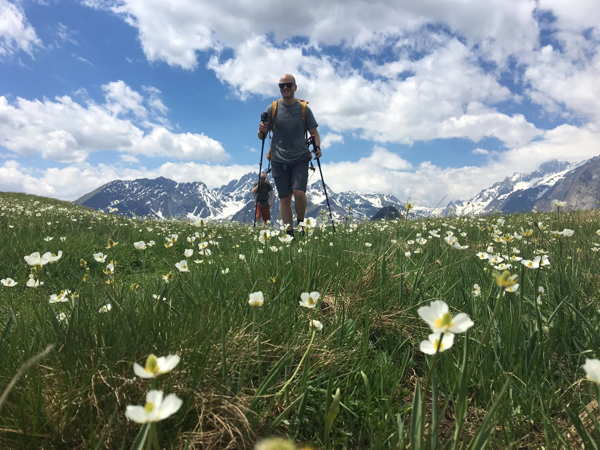 En turgåer med vandrestaver går gjennom en grønn alpin eng fylt med hvite villblomster, med snødekte fjelltopper som hever seg under en blå, skyete himmel i bakgrunnen.